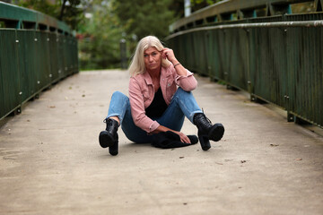 Woman blonde in her fifties with a pink denim jacket, blue jeans with a ripped black sweater and a black scarf, portrait on a bridge.