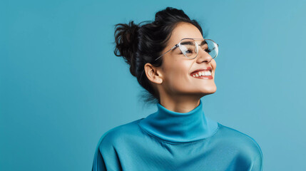 Horizontal photograph of a self embracing and joyful woman who appears optimistic but shy donning transparent glasses and a casual turtleneck standing alone against a blue background 