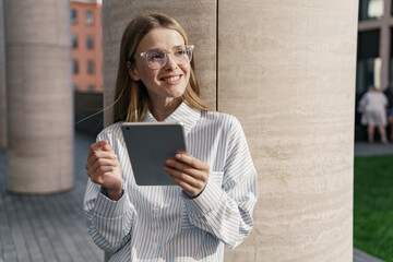Smiling young woman in glasses using a tablet on a sunny city street.