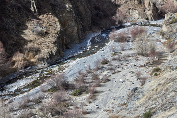 Small mountain river of beautiful color with stones banks and rocky slope. Branches of trees and bushes without leaves. Autumn natural landscape, top view. Nature Kyrgyzstan