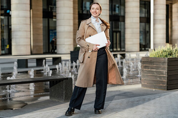 Fototapeta premium Chic businesswoman in trench coat with documents strides confidently outside corporate building.
