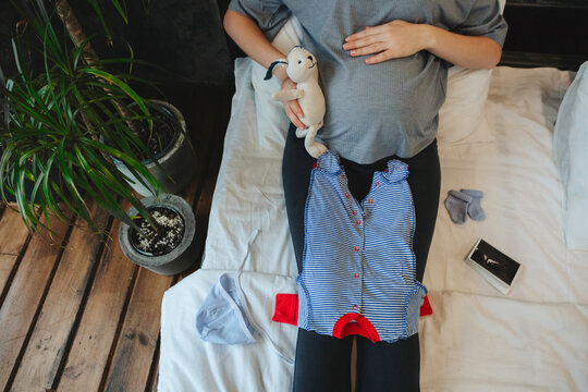 Pregnant woman looking at adorable newborn clothing while relaxing at home, preparing for childbirth