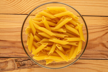 Raw pasta, penne, in glass plate on wooden table, macro, top view.