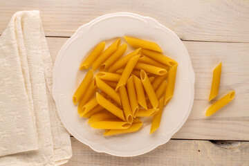 Raw pasta, penne, in a ceramic plate on a wooden table, macro, top view.