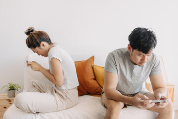 Asian Thai couple sitting back to back on sofa at home, Both man and woman using mobile phone, playing game online, ignoring living in apartment room.