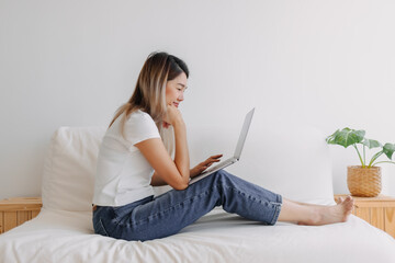 Happy Asian Thai woman sitting on sofa and putting computer on her knees, working at room apartment alone, using laptop for working.