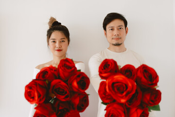 Asian Thai couple lover in white holding red roses, standing over white background wall, Both giving flowers and looking at camera minimal.