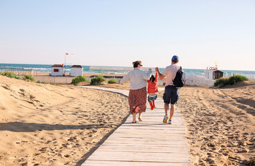 Happy family, mother father and little daughter, walking towards summer sea beach along wooden path