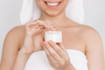 Obraz na płótnie Canvas Cropped shot of a young smiling woman in a white towel after shower taking moisturizing cream with the finger holding cosmetic jar in her hand on a light background. Skin care. Cosmetology and beauty