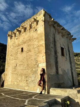 Vintage pin-up, donned in black and red, poses by a medieval tower on Gargano with a bold slit and captivating smile, enhancing the allure amid the historic setting.