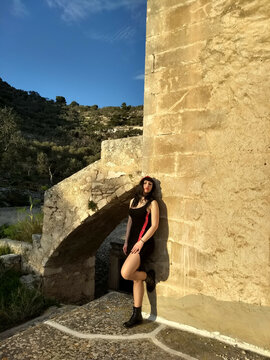 Vintage pin-up, donned in black and red, poses by a medieval tower on Gargano with a bold slit and captivating smile, enhancing the allure amid the historic setting.