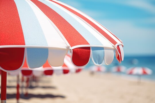 Close up image of a red blue and white vertical striped beach umbrella on a background of a beach with sun loungers and blue sea
