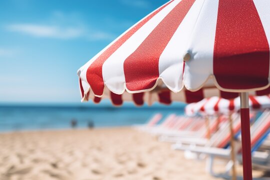 Close up image of a red and white vertical striped beach umbrella on a background of a beach with sun loungers and blue sea