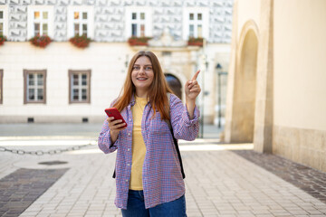 Fototapeta premium Picture of pretty young woman with backpack staying on the street. 30s tourist walking with phone and pointing finger on old city street. Use technology concept, Traveling Europe in summer