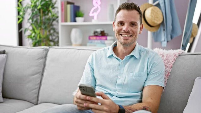 A relaxed hispanic man with a beard enjoys using a smartphone in his modern living room, signifying connectivity and leisure at home.