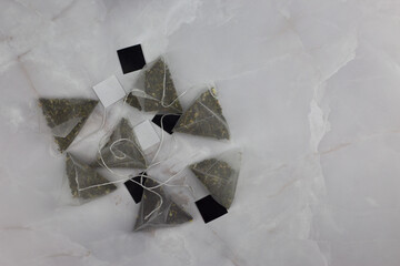 Green tea bags with fruits in the shape of pyramids lie on a white marble table. Transparent nylon tea bag with string and cardboard for holding by hand while brewing the drink - obrazy, fototapety, plakaty