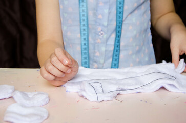 The hands of a handmaiden girl, a child in a blue blouse draws a cut-out with a pencil and then sews a cat toy out of white fabric. 