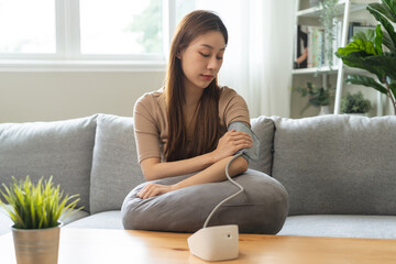 Hypotension, asian young woman sitting on couch checking blood high pressure and heart rate with digital pressure monitor machine making self check up on her arm at home. Healthcare and medical. - obrazy, fototapety, plakaty