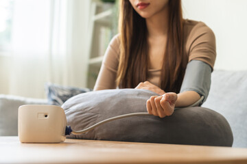 Hypotension, closeup of hand asian young woman sitting on couch checking blood high pressure and heart rate with digital monitor machine making self check up on arm at home. Healthcare and medical.