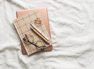 Notepad, tablet, hair clip, glasses, pens on the white sheets of the bed. Cozy morning, planning concept