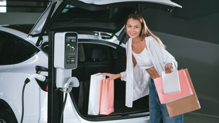 Young woman travel with EV electric car to shopping center parking lot charging in downtown city...