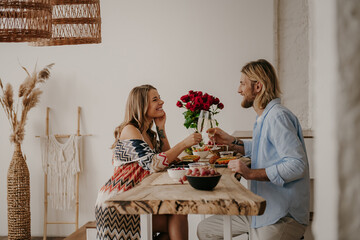 Joyful young loving couple toasting with champagne while having romantic dinner at boho style home