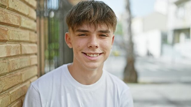 A smiling young caucasian male teenager in a white shirt standing on a sunny urban street.