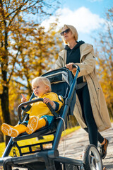 Young beautiful mother wearing a rain coat pushing stroller with her little baby boy child, walking...