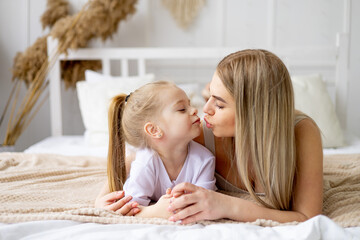 mom and daughter play, hug and kiss at home on the bed, lifestyle, tender relationship of a young...