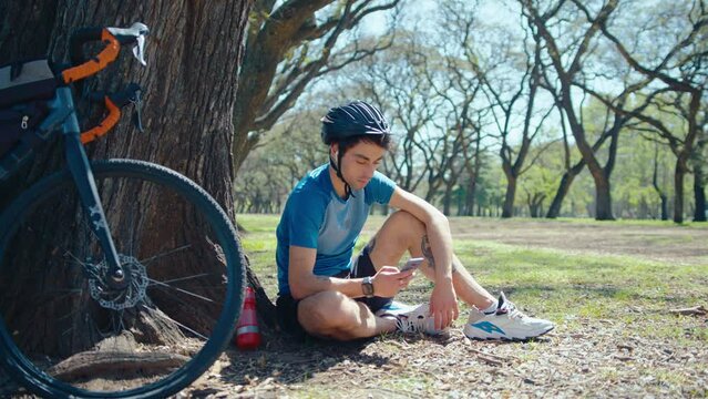 Young male cyclist in sports clothing and bike helmet sitting on the ground by tree and texting on mobile phone during break in the park