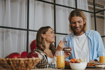 Smiling young woman feeding her boyfriend with breakfast while enjoying healthy food at the kitchen
