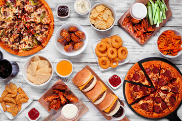 Junk food table scene. Pizza, hamburgers, chicken wings and salty snacks. Top down view over a...