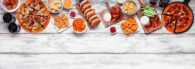 Junk food top border. Pizza, hamburgers, chicken wings and salty snacks. Above view over a white...