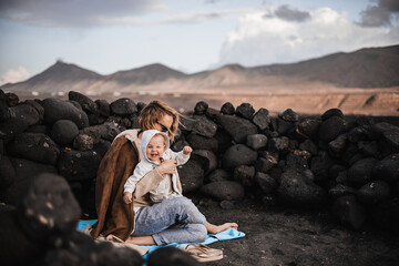 Mother enjoying winter vacations playing with his infant baby boy son on black sandy volcanic beach...