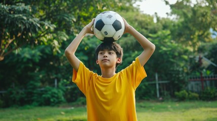 Teenage boy raises soccer ball over head dreams of becoming professional football player and playing in Major League. Young guy in yellow T-shirt stands on green lawn