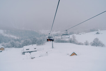 Fototapeta na wymiar Skiers ride a chairlift up the mountain to snow-covered cottages among a coniferous forest