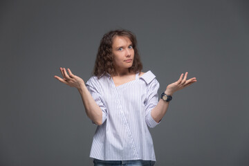 A young woman on a gray background with bewilderment on her face spreads her hands and shrugs her shoulders.