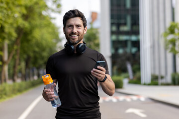 Portrait of a smiling young man running on the city street, standing in headphones, holding a...