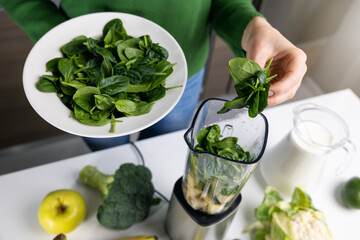 woman puts fresh green spinach in blender to make healthy detox smoothie from vegetables and fruits...