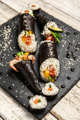 Slate board with tasty sushi rolls, cones, sauce and sesame seeds on light wooden background, closeup