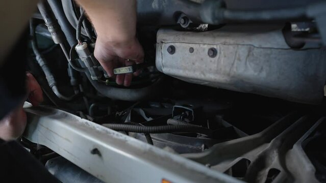 Hands of a mechanic working on a car engine, connecting a hose with a clip. The engine compartment is open, showing various components. The image conveys auto repair and maintenance in progress.