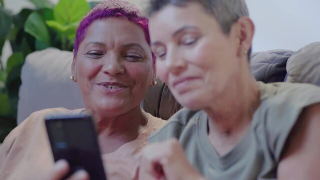 Young happy senior lesbian couple having fun using smartphone at home. Looking at mobile phone buying retail products shopping online, ordering delivery, checking social media apps.
