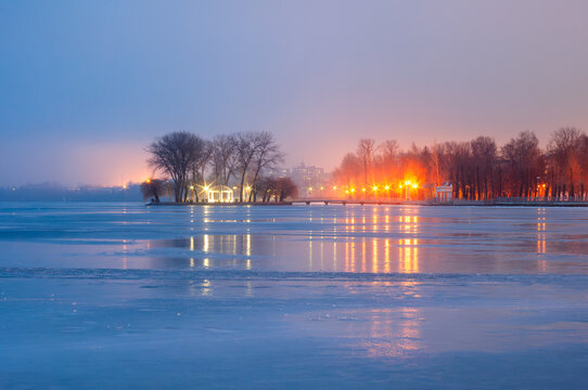 Lights of evening city lanterns. View of the embankment and the bridge to the island from the surface of the frozen lake. Island of Love and Shevchenko Park in Ternopil, Ukraine