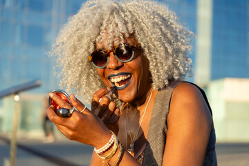 Afro-american woman with grey afro hair putting on lipstick in urban environment.