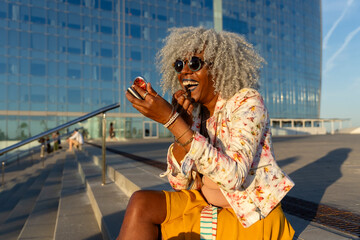 Afro-american woman with grey afro hair putting on lipstick in urban environment.