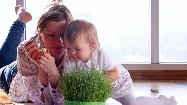 Woman with a toddler playing and coloring eggs, preparing for easter at home. Kid putting small colored eggs in the grass, egg hunt at home with an infant