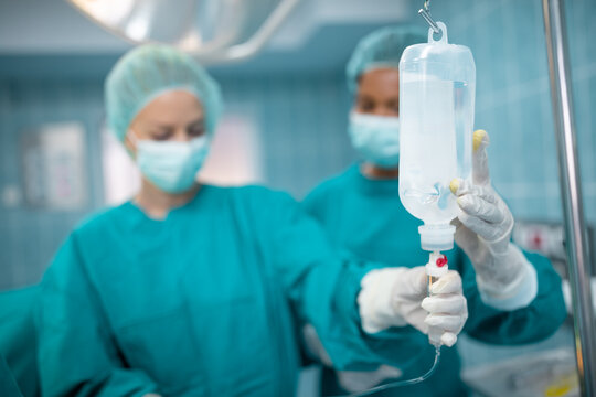 Closeup of IV drip in operating room. Female surgeon and nurse preparing intravenous drip for patient in operating theatre. Focus is on the IV drip.