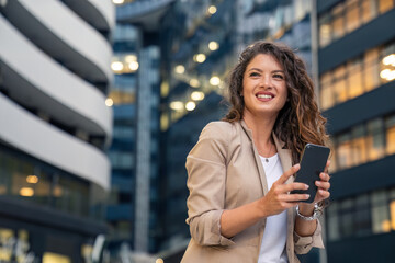 Young adult smiling professional business woman wearing suit holding smartphone and looking away...