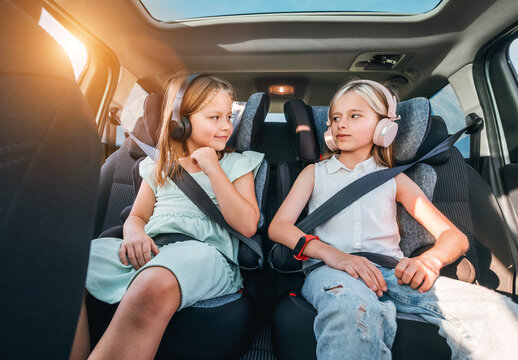 Portrait of two positive smiling sisters sitting in child car seats fastening safety belts listening to music using headphones. Happy childhood, auto journey concept image.