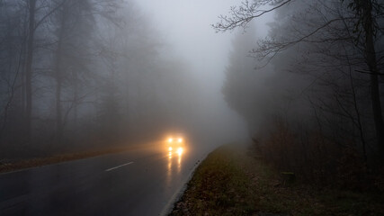 Car with the illuminated headlights on the wet road in the dark and foggy forest